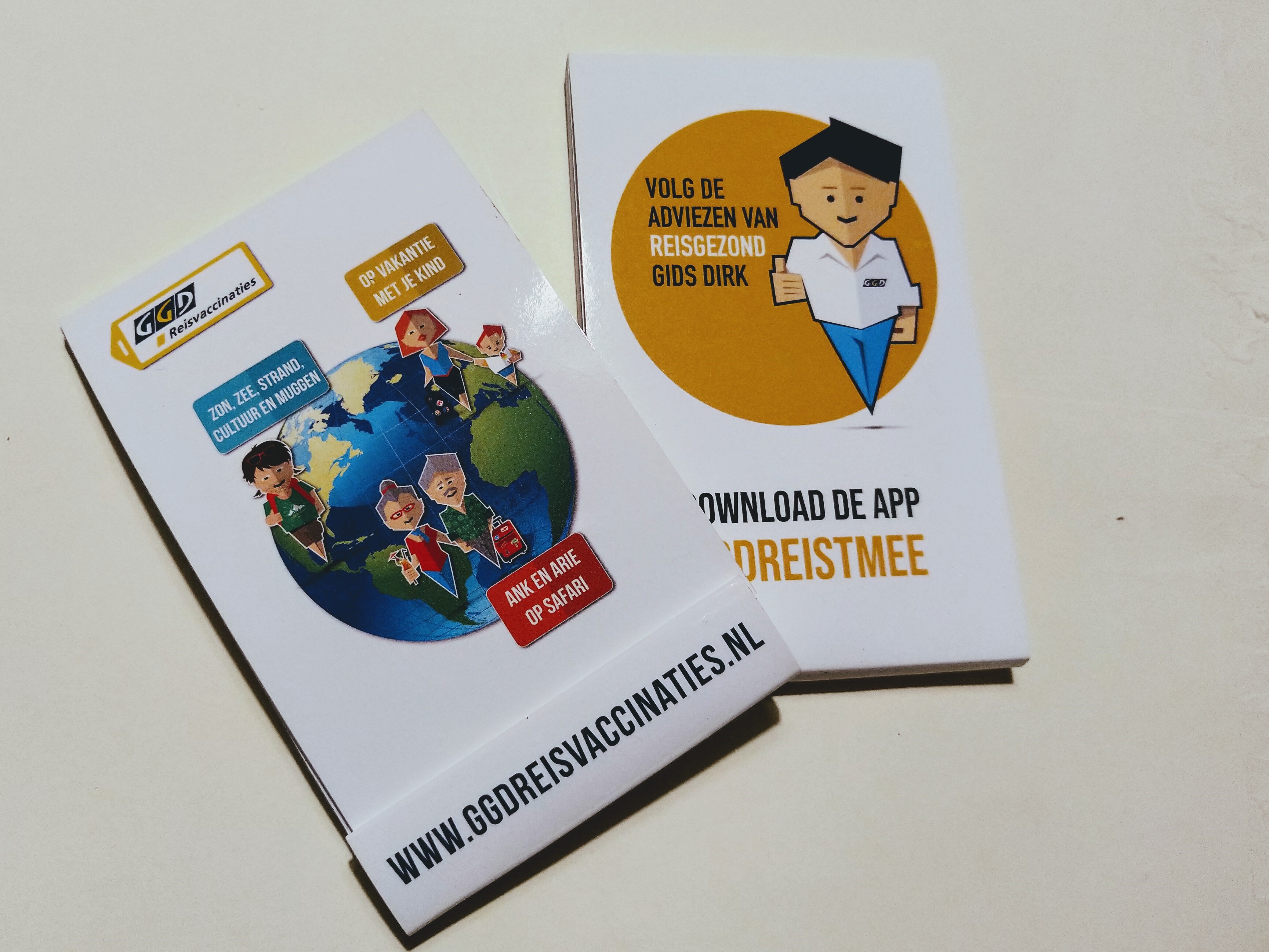 Wit notitieblokje met logo GGDReisvaccinaties. Op voorkant wereldbol met avatar jonge backpacker: zon, zee, strand, cultuur en muggen. Avatar: moeder en kind: op vakantie met je kind. Avatar twee fitte vijftigplussers: Ank en Arie op safari. Onderaan: www.ggdreisvaccinaties.nl Op achterkant gele cirkel met avatar Dirk: volg de adviezen van Reis Gezond Gids Dirk. Download de app ggdreistmee.