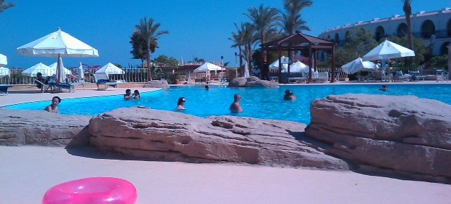 Zwembad op resort in Hurghada Egypte. Voor het zwembad ligt een roze opblaasband. Naast het zwembad staal witte parasols. In het zwembad zwemmen mensen.