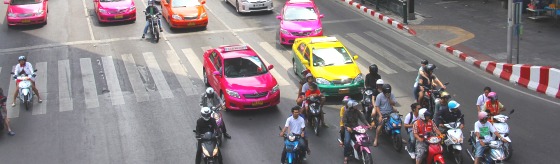Foto van kruispunt in Bangkok. Op de weg staan een roze, gele en oranje taxi auto's. Daarvoor staan scooters en motoren met daarop personen. 