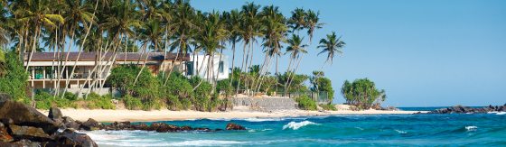 Baai in Sri Lanka. Helder blauw water en een wit strand. Op het strand staan palmbomen en in de verte een gebouw.