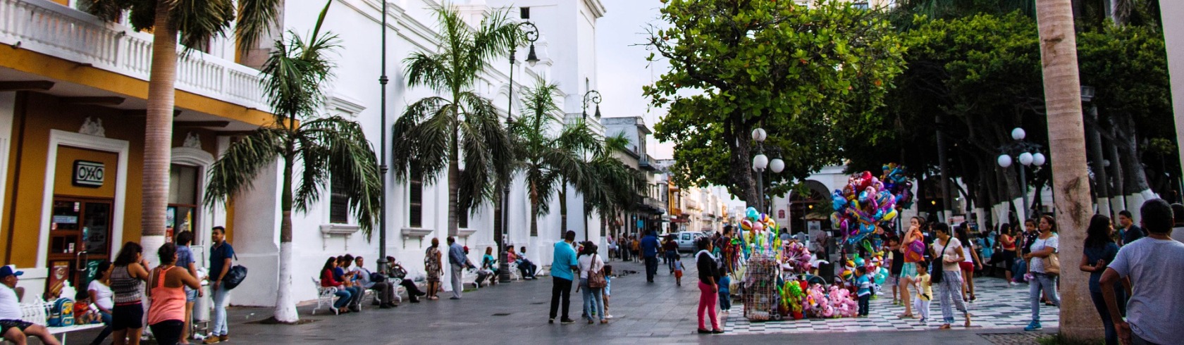 Plein in een stad in Mexico. Lopende mensen, palmbomen tegen wit gebouw, kraam met ballonnen. 