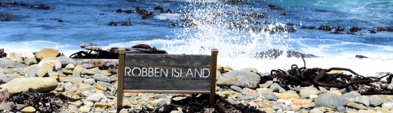 Foto van zeegolven die tegen de stenen omhoog botsen. Op de stenen staat een bord: Robben Island.