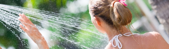 Vrouw staat buiten onder een straal van de douche. Vrouw draagt een bikini top.