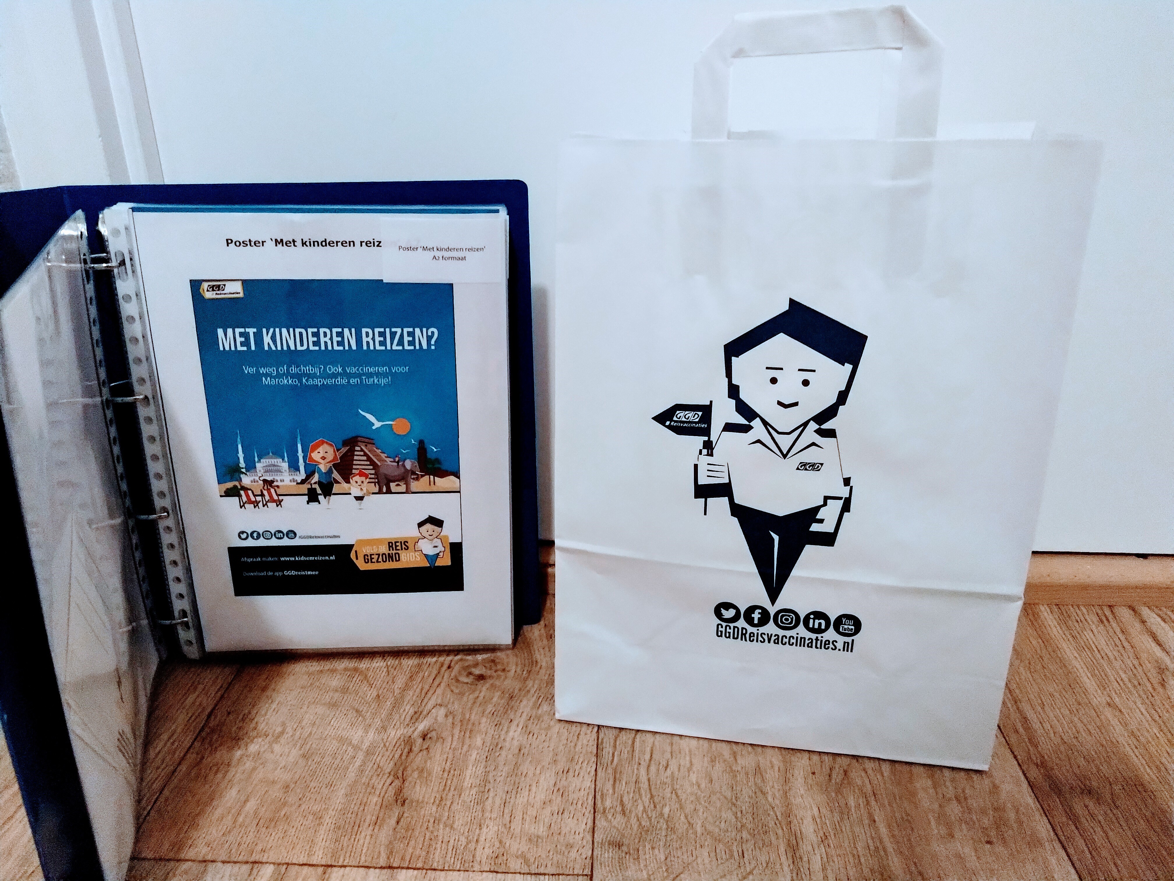 Wit papieren draagtas met avatar Dirk en logo van facebook, instagram, twitter, linked in en YouTube. ggdreisvaccinaties.nl Daarnaast staat een blauwe map met daarin poster: met kinderen op reis.