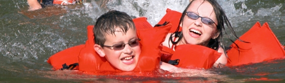 Twee zwemmende kinderen met rode zwemvesten aan het spelen in het water. Meisje en jongen dragen een zonnebril.
