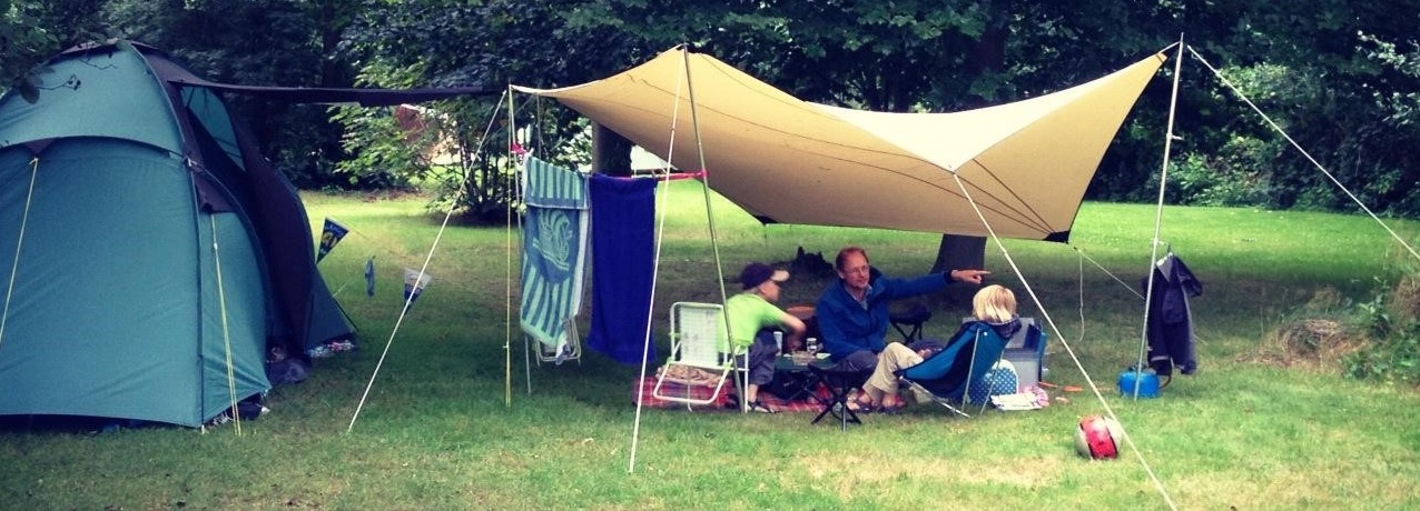 Tent en tentzeil op een camping in het bos. Twee mensen zitten bij de tent. 